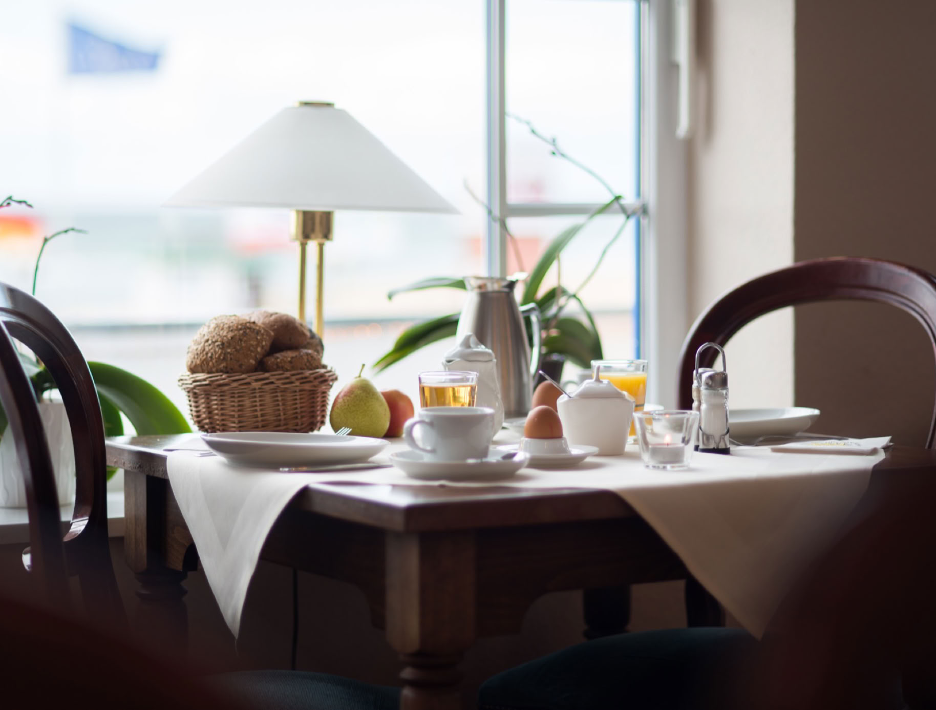 Ein gedeckter Tisch mit einer weißen Tischdecke, Geschirr, einer Lampe, einem Brotkorb und Pflanzen im Hintergrund.