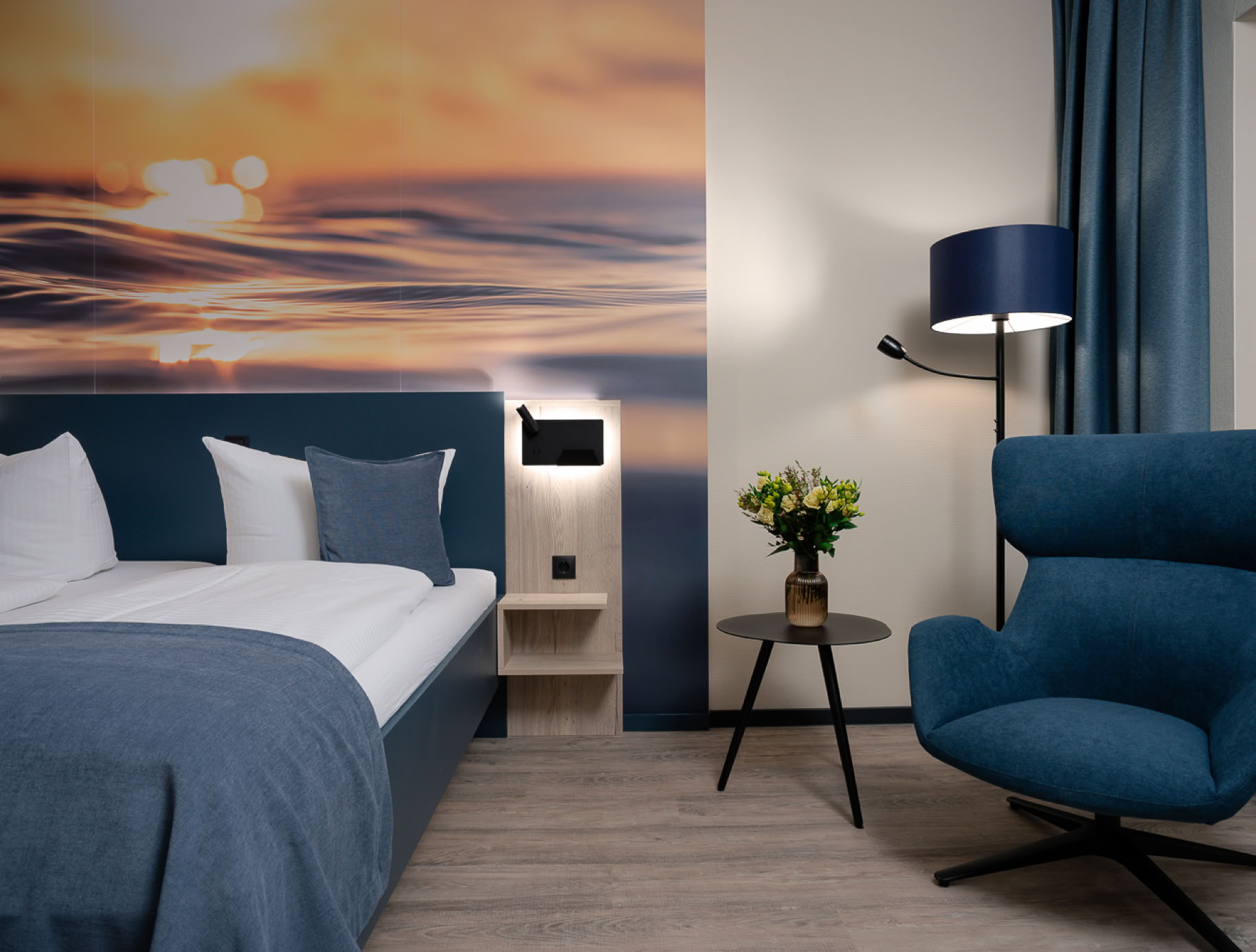 Ein Hotelzimmer mit einem Doppelbett, blauer Decke, Holzboden, Nachttischlampen, einem Stuhl und einem Fenster mit Vorhängen.